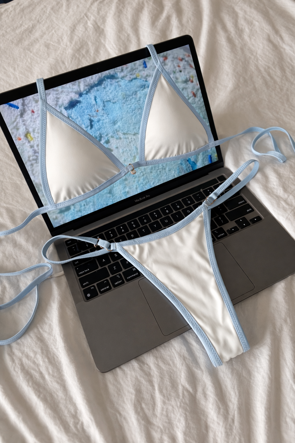 White triangle bikini top and matching thong bottoms with light blue trim displayed on a laptop keyboard on white fabric background