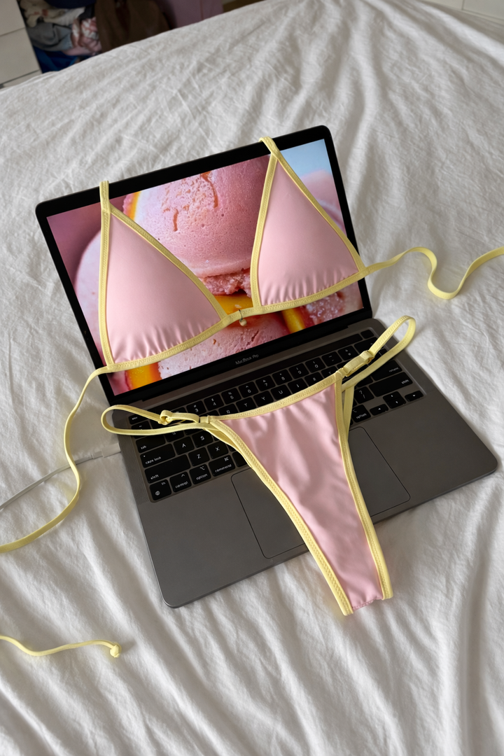 Pastel pink triangle bikini top and matching thong with yellow trim laid on open laptop keyboard on white sheets
