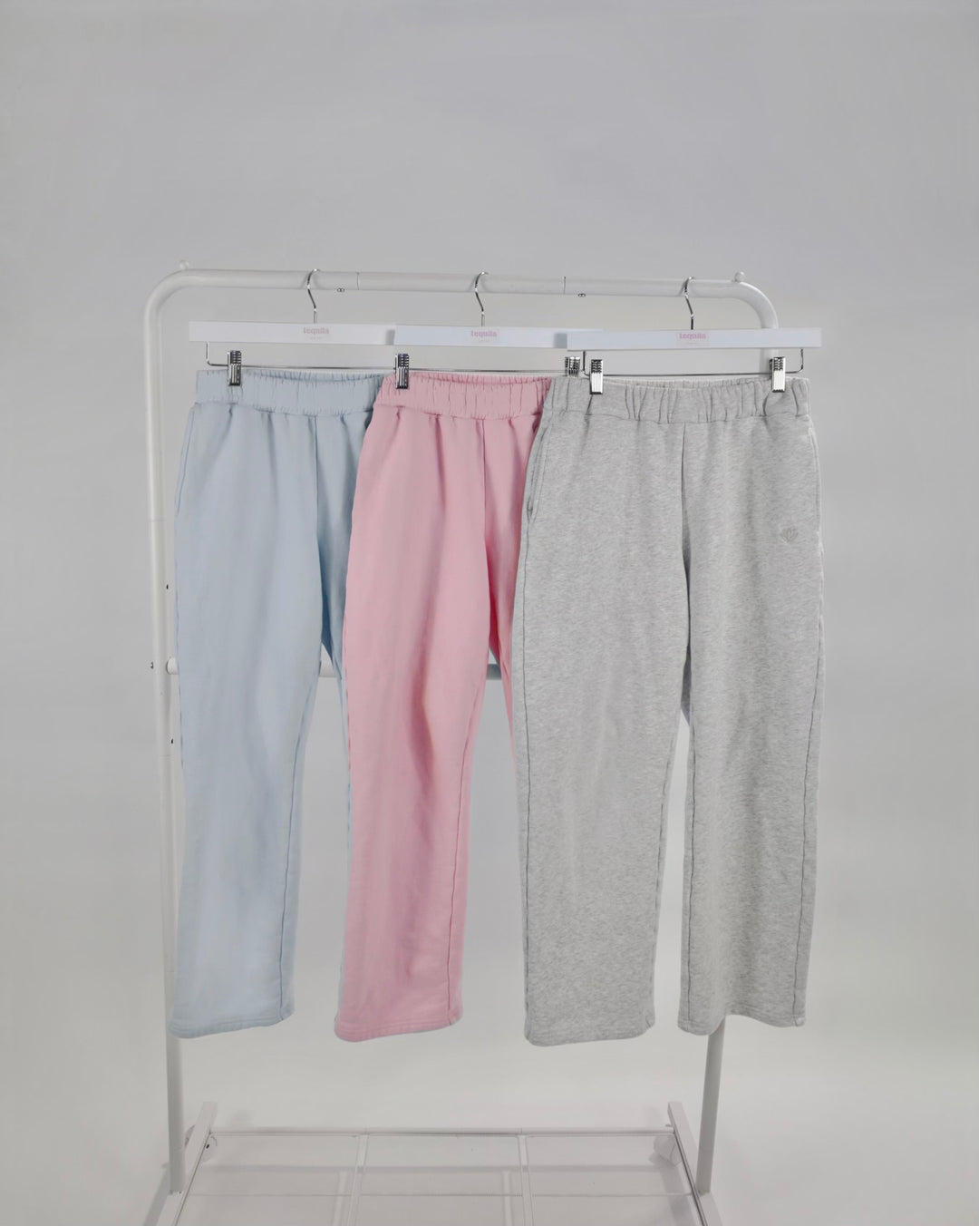 Three pairs of Bunny Sweatpants in light blue, pink, and gray hanging on a white clothing rack against a gray background