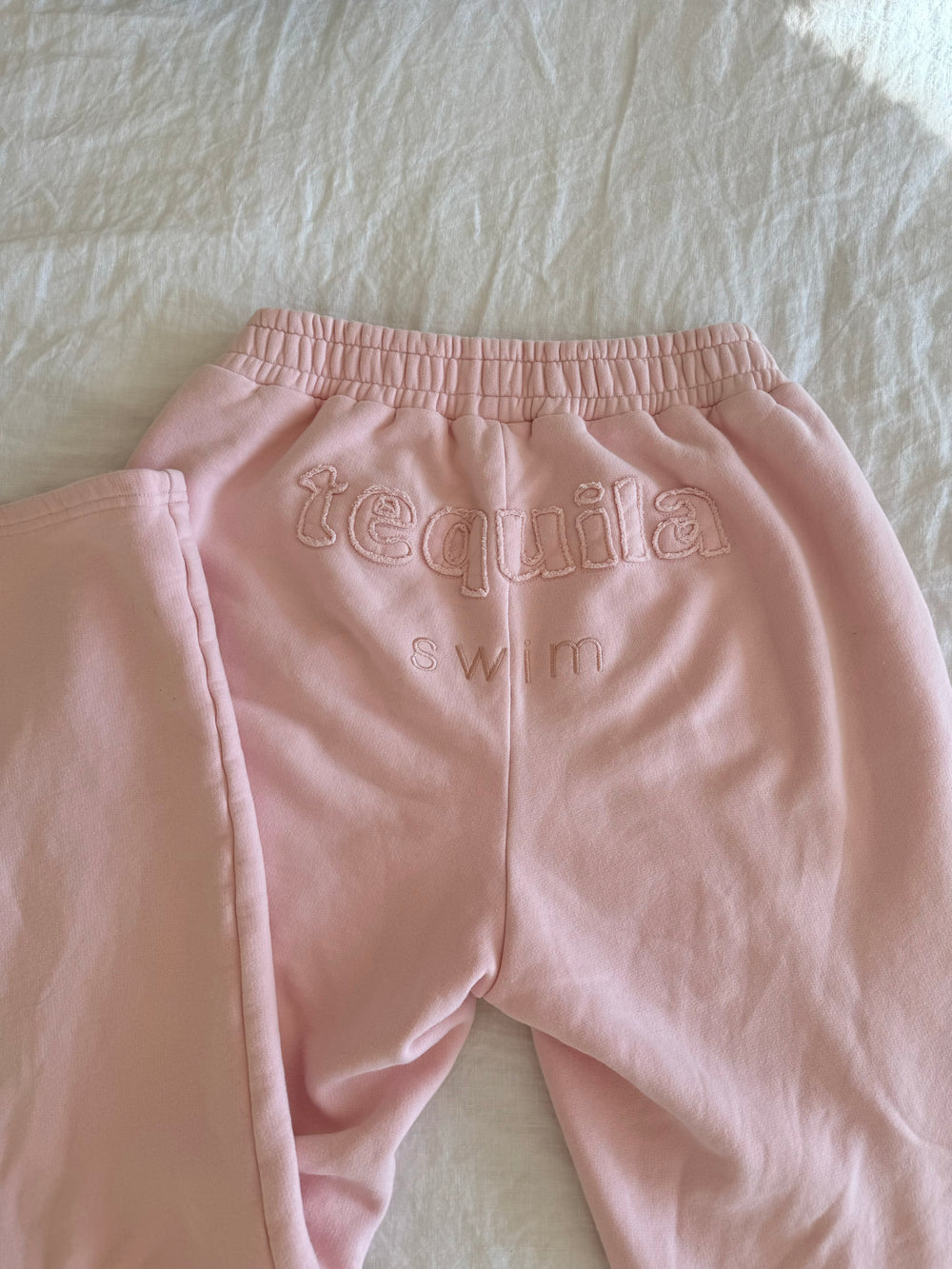 Pink Bunny Sweatpants with embroidered "tequila swim" logo on back, laid flat on white fabric background.