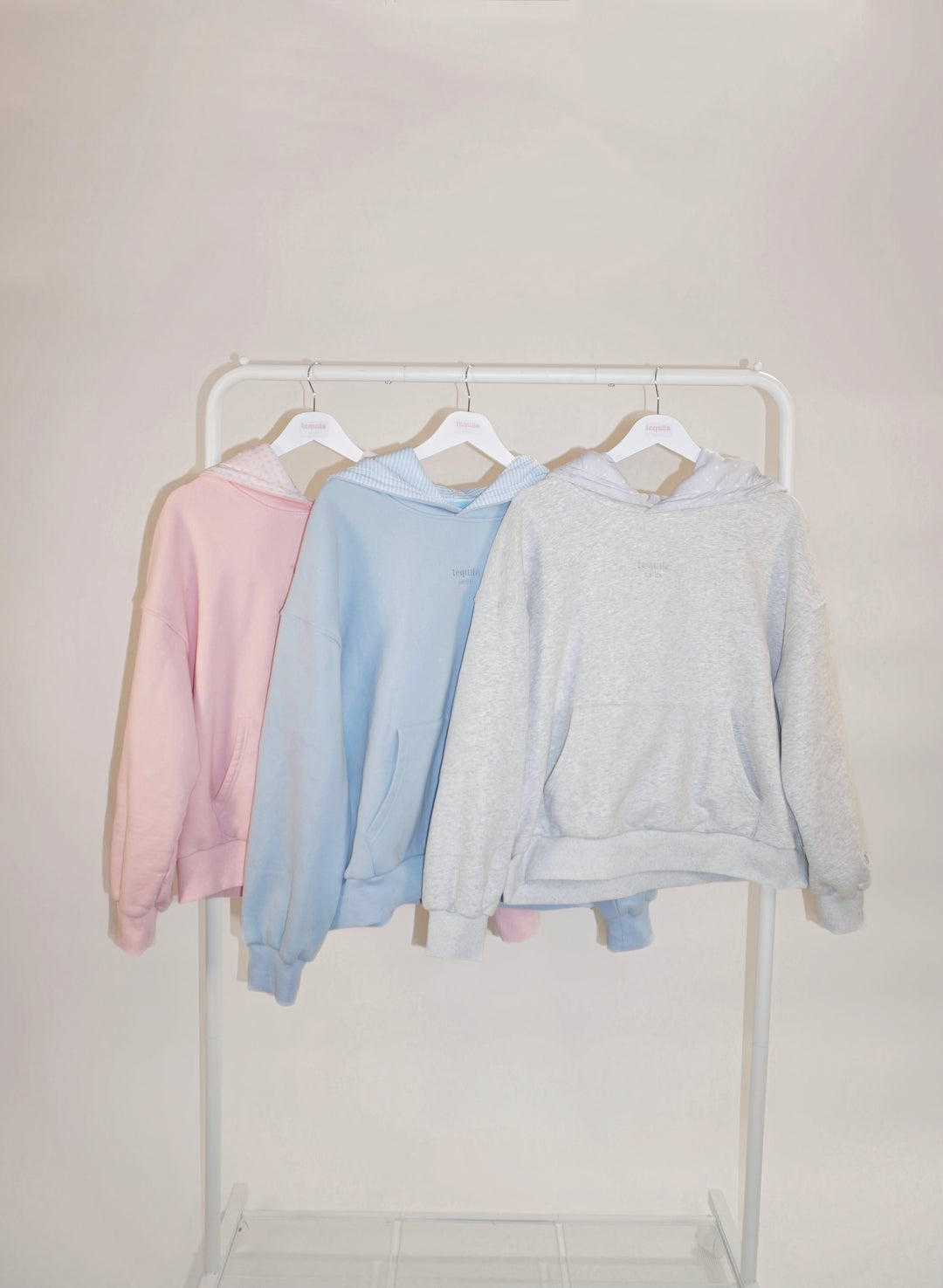 Three pastel-colored Bunny Hug Hoodies in pink, blue, and gray hanging on a white rack against a light background