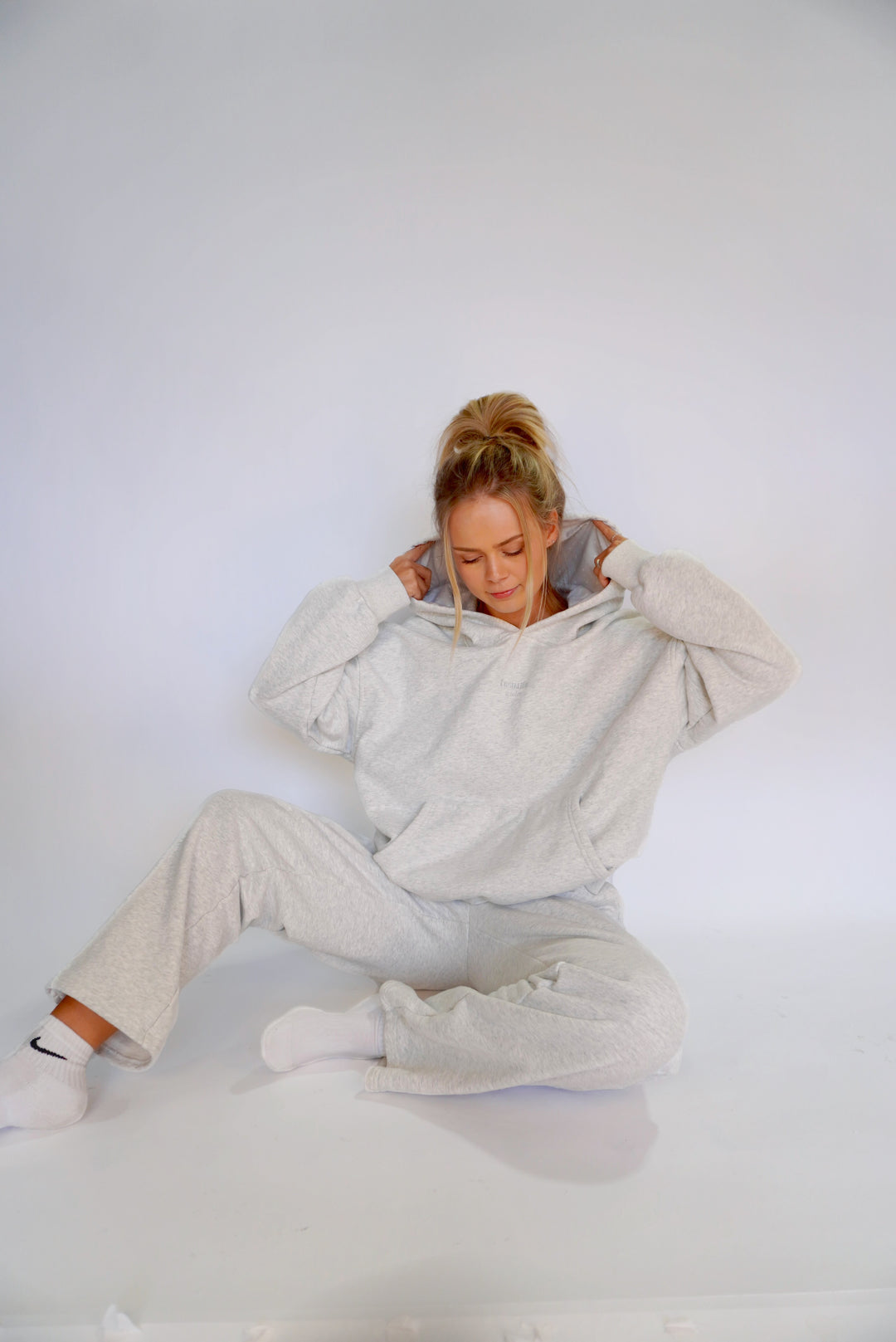 Woman wearing light gray Bunny Hug Hoodie and matching wide-leg pants sitting on the floor against a plain background