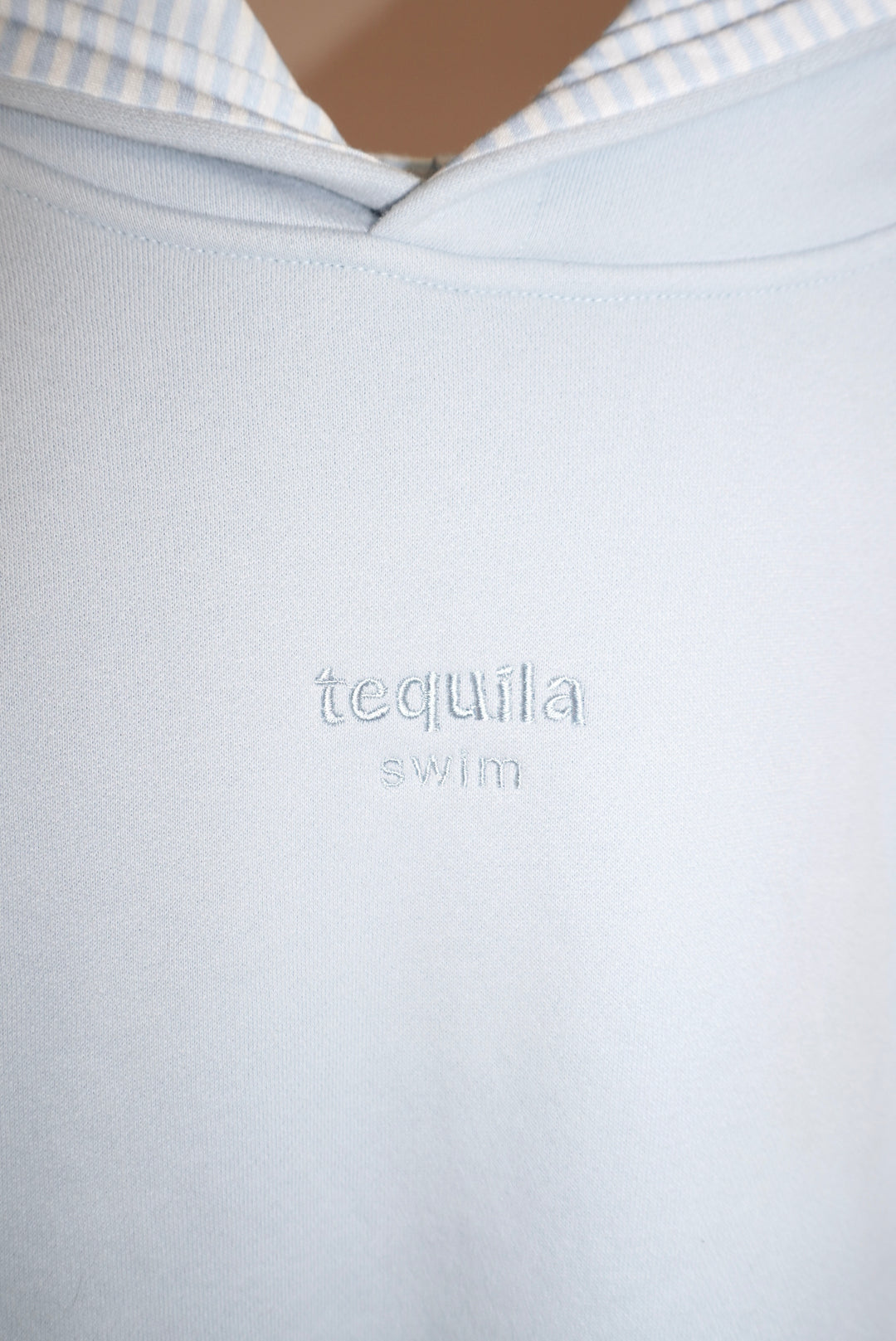 Close-up of light blue Bunny Hug Hoodie with embroidered "tequila swim" logo on chest and striped inner hood lining