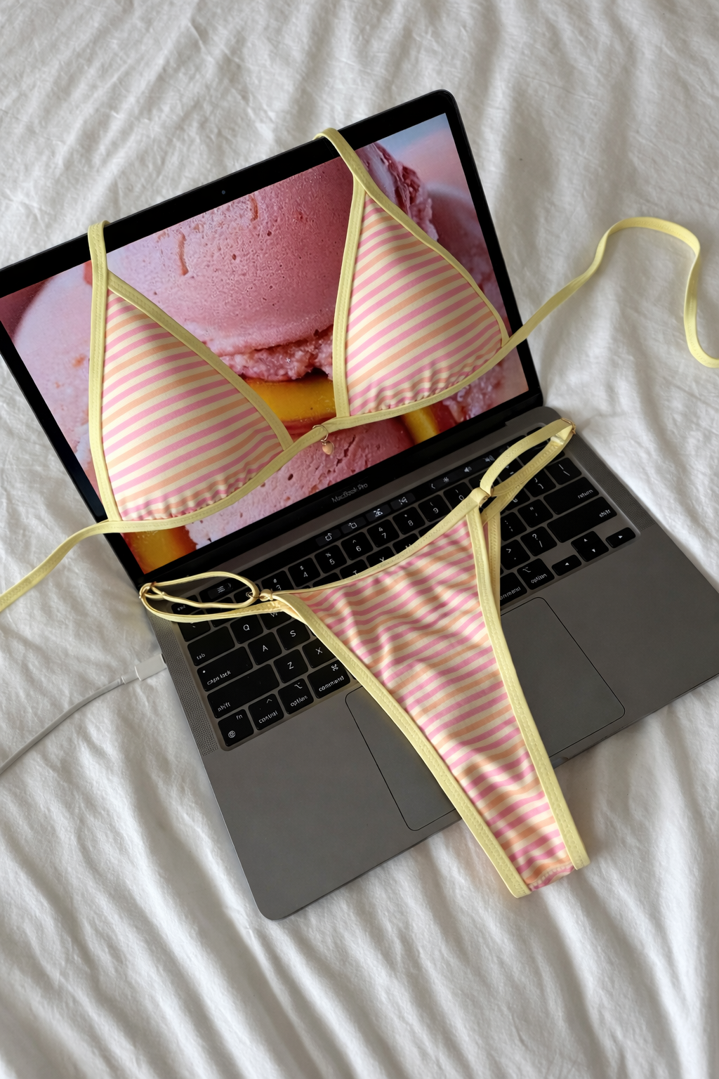 Pink and white striped triangle bikini top and matching bottoms with yellow trim laid on open laptop keyboard