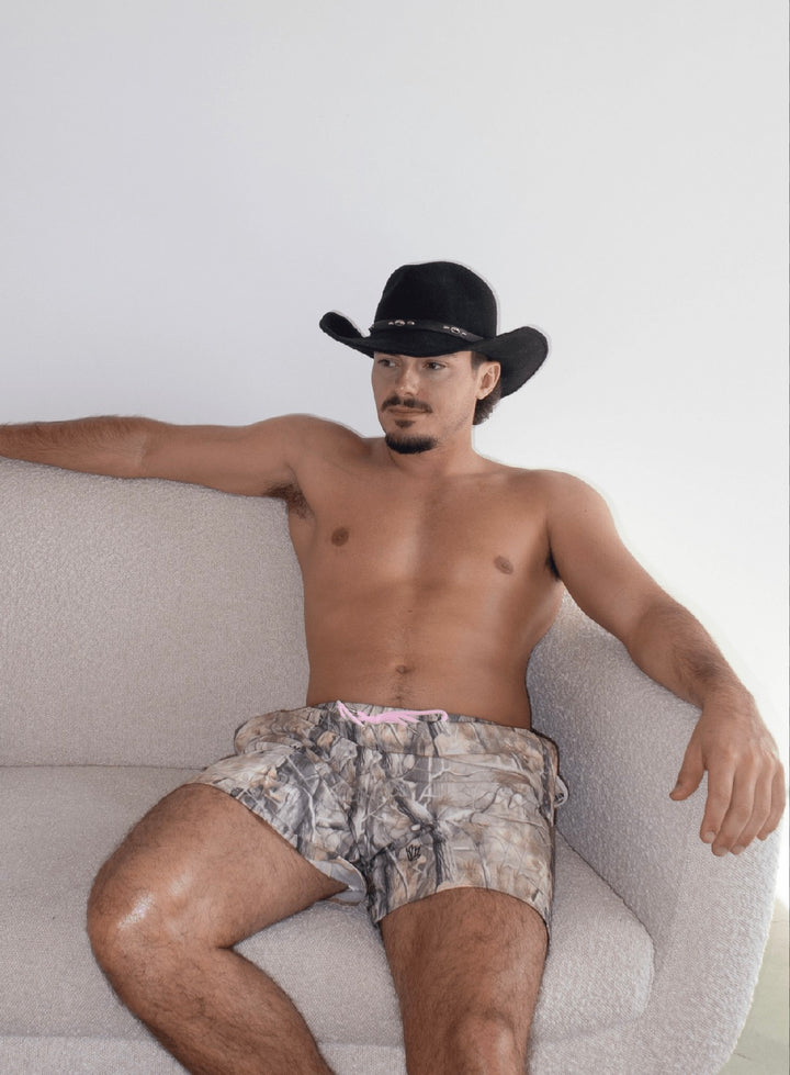 Man wearing camo Jaimo 5.5" shorts and black cowboy hat sitting on a beige couch with relaxed pose