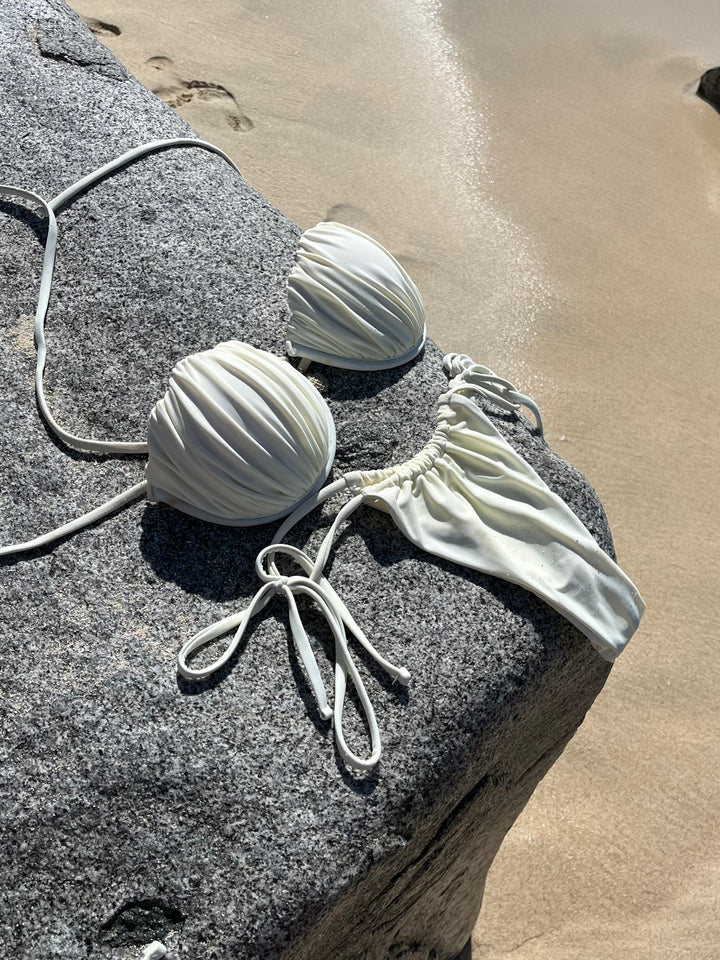White Ariel shell ruched bikini top and matching bottoms with tie straps on a beach rock near sand.