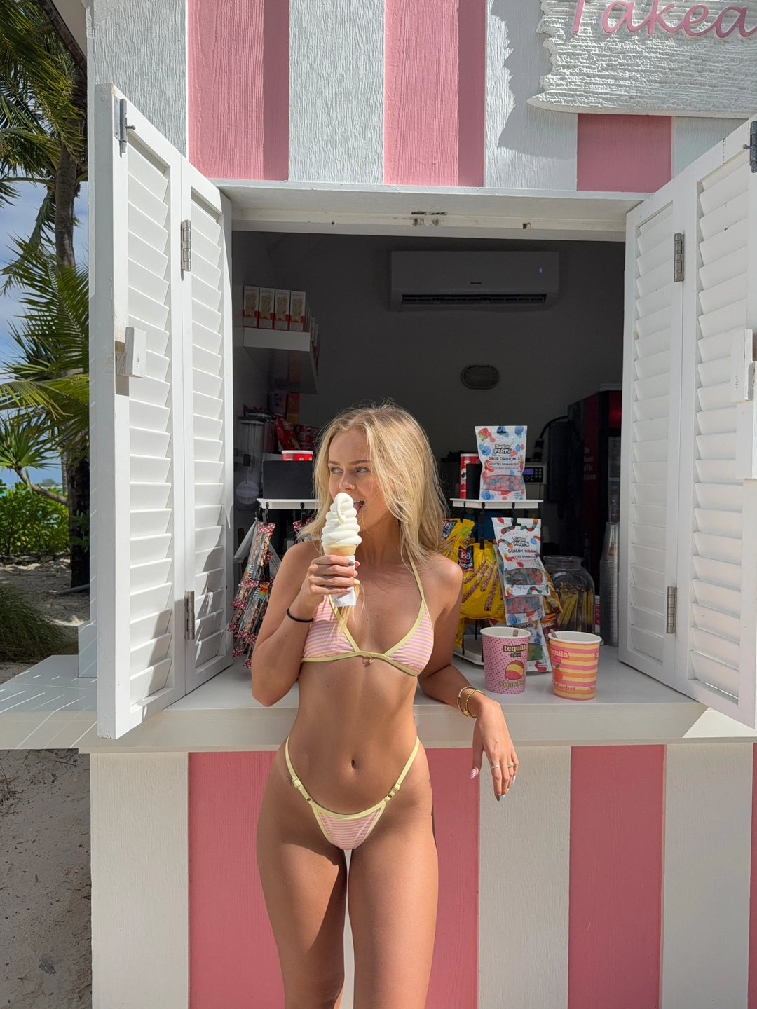 Woman in pink and yellow striped bikini standing at a pink and white striped ice cream shack holding a soft serve cone