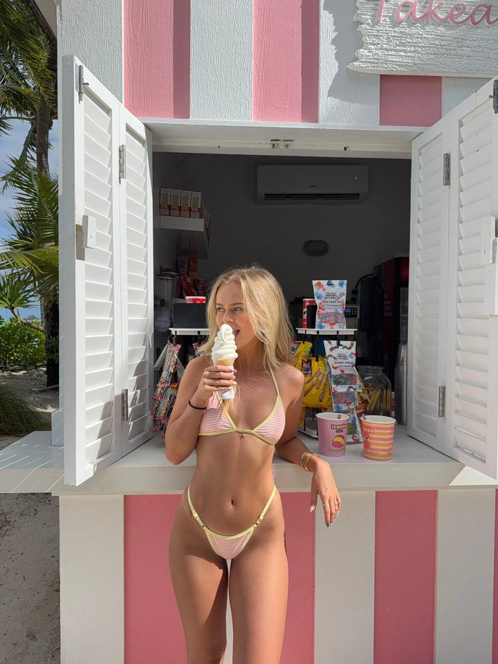 Woman in pink and yellow striped bikini standing at a pink and white striped ice cream shack holding a soft serve cone