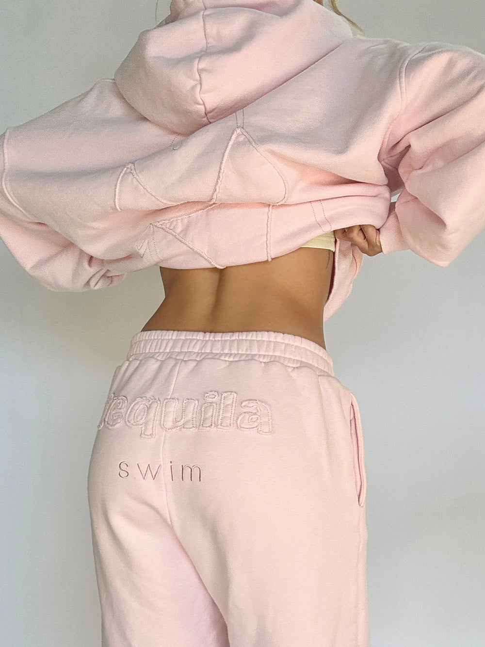 Woman lifting light pink hoodie to reveal matching sweatpants with "Tequila swim" embroidered on back waistband